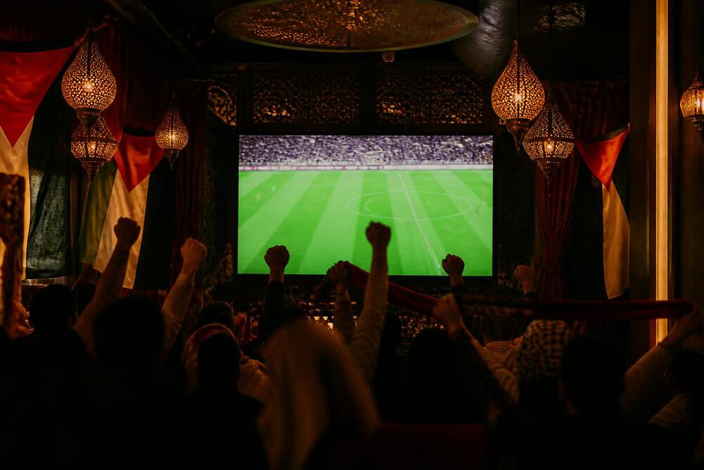 Supporters watching a soccer match in a lantern-lit lounge with Palestinian flags